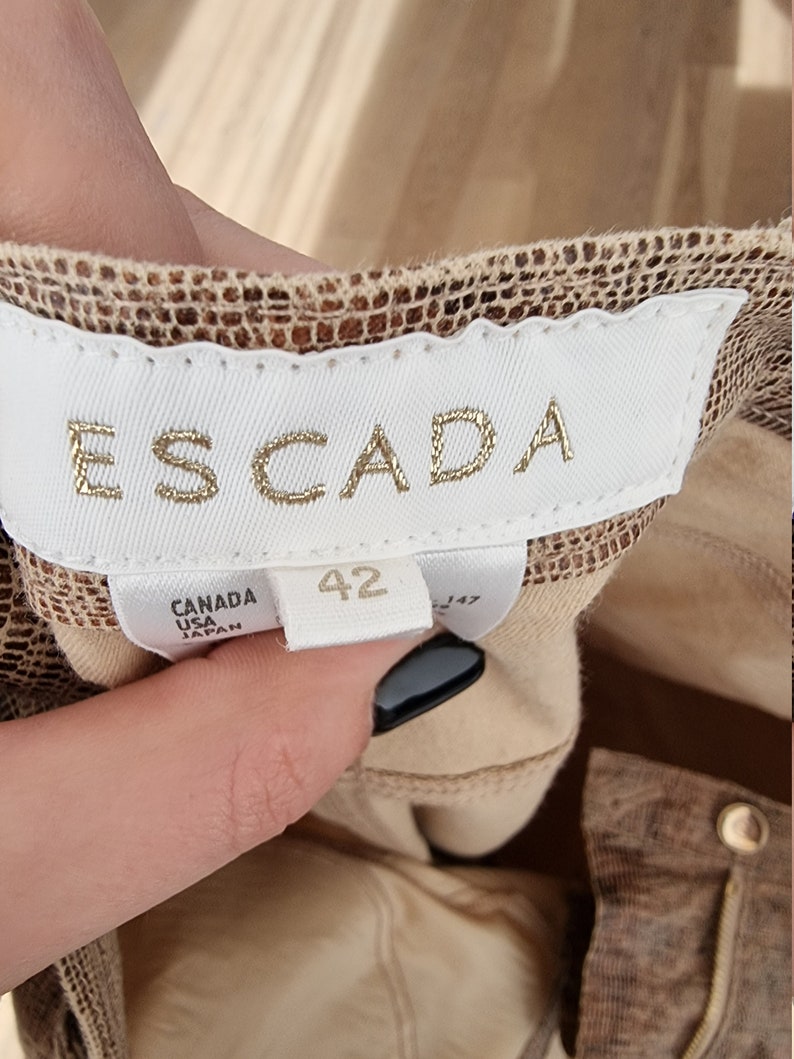 May include: Close-up of a clothing label with the brand name "ESCADA" in gold lettering. The label is white with the size "42" printed on it. The label also says "CANADA USA JAPAN". The fabric is a brown and tan snakeskin pattern.