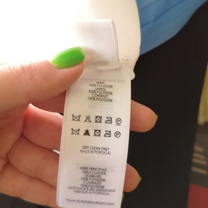 May include: White care label with black text and symbols. The label states "Main: 100% Polyester, Lining: 100% Polyester, Contrast: 100% Polyester". The label also includes instructions for dry cleaning only and states "Made in Portugal".