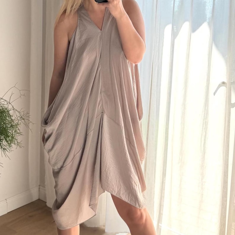 Puede incluir: Vestido sin mangas de color topo con escote en V y dise&ntilde;o asim&eacute;trico drapeado. El vestido tiene un ajuste holgado y relajado y parece estar hecho de una tela suave y sedosa. El vestido llega a la rodilla y es adecuado para ocasiones informales o semiformales.