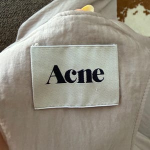 Puede incluir: Primer plano de una prenda beige claro con una etiqueta rectangular. La etiqueta, cosida a la tela, muestra la palabra "Acne" en una fuente negra y en negrita. La tela parece suave y ligeramente arrugada.