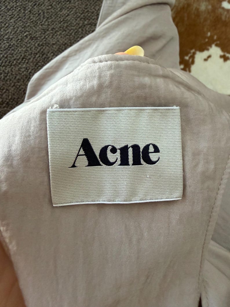 Puede incluir: Primer plano de una prenda beige claro con una etiqueta rectangular. La etiqueta, cosida a la tela, muestra la palabra "Acne" en una fuente negra y en negrita. La tela parece suave y ligeramente arrugada.