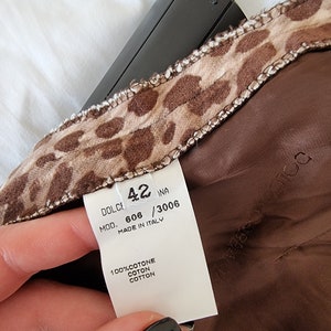 Puede incluir: Una etiqueta blanca con texto negro que dice "DOLCE & GABBANA MOD. 606 / 3006 MADE IN ITALY 100% COTONE COTON COTTON" con un tejido de estampado de leopardo marr&oacute;n y beige detr&aacute;s. La etiqueta est&aacute; unida a una prenda marr&oacute;n.