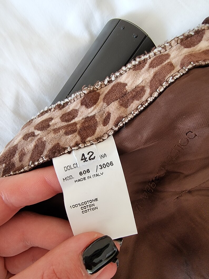 Puede incluir: Una etiqueta blanca con texto negro que dice "DOLCE & GABBANA MOD. 606 / 3006 MADE IN ITALY 100% COTONE COTON COTTON" con un tejido de estampado de leopardo marr&oacute;n y beige detr&aacute;s. La etiqueta est&aacute; unida a una prenda marr&oacute;n.