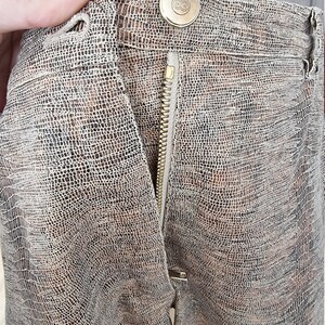 May include: Close-up of a pair of brown and tan patterned pants. The pants have a zipper and a button closure. The button is gold-colored with an embossed design. The fabric has a textured, reptile-like pattern.