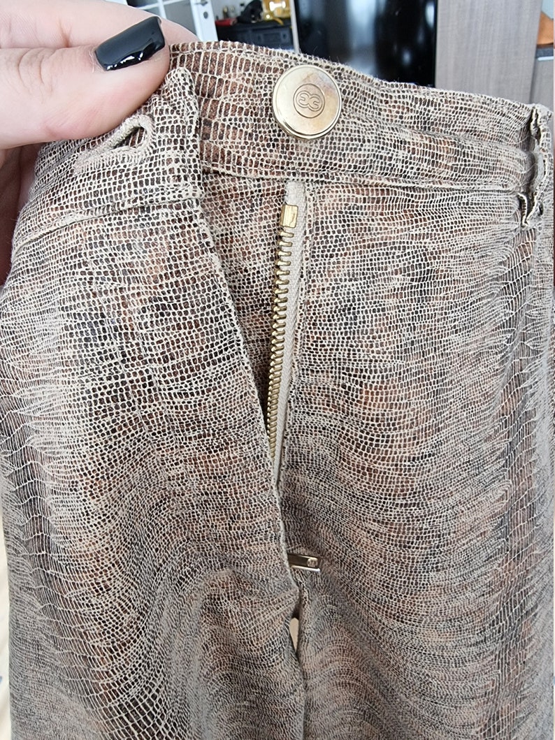 May include: Close-up of a pair of brown and tan patterned pants. The pants have a zipper and a button closure. The button is gold-colored with an embossed design. The fabric has a textured, reptile-like pattern.
