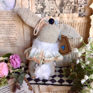 May include: A handmade white rabbit doll with a white fur tail and a lace skirt. The doll is wearing a blue and white heart tag that says "White Rabbit". The doll is sitting on a black and white checkered surface.