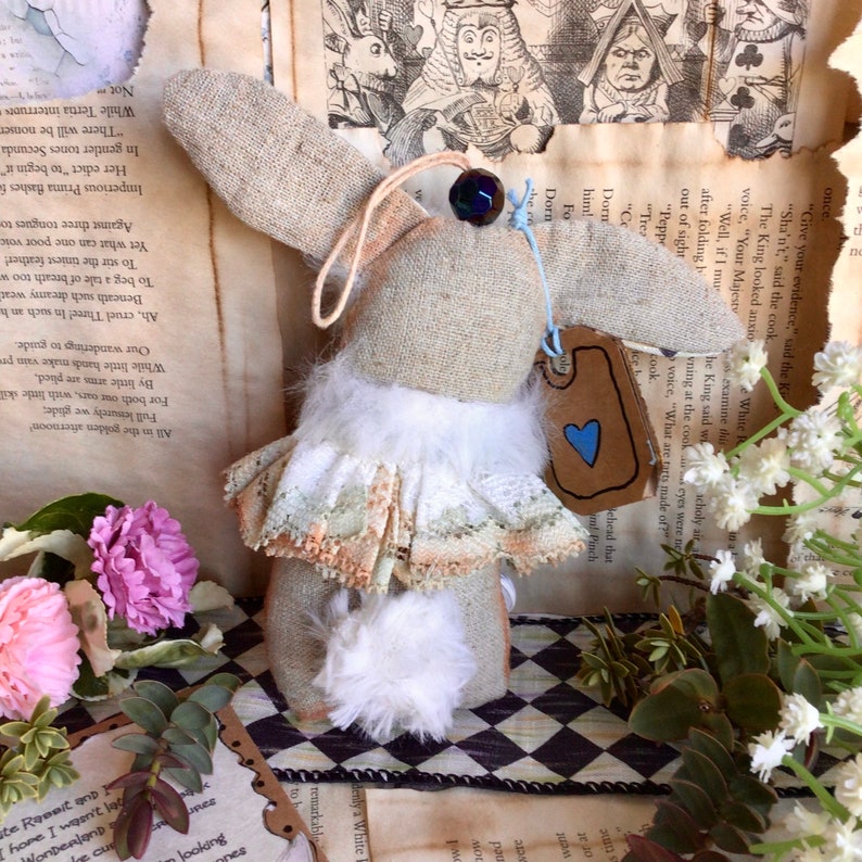 May include: A handmade white rabbit doll with a white fur tail and a lace skirt. The doll is wearing a blue and white heart tag that says "White Rabbit". The doll is sitting on a black and white checkered surface.