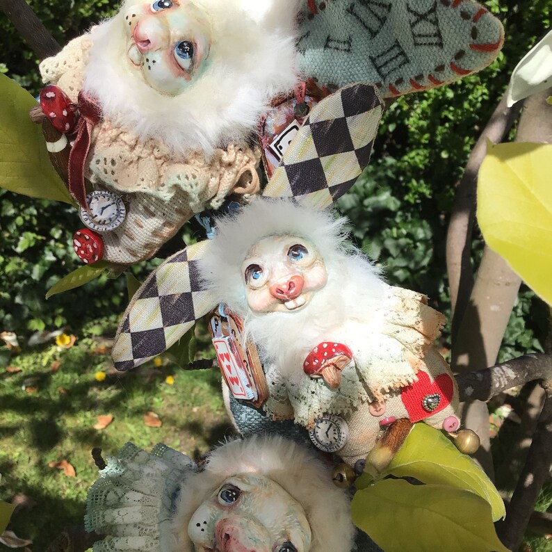 May include: Three white rabbit plushies with painted faces and detailed clothing. The plushies are decorated with lace, fabric flowers, and a checkered pattern. One plush is holding a mushroom.