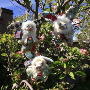 May include: Three white rabbit plushies with different fabric textures and designs. Each plush has a different face and is decorated with fabric flowers, hearts, and playing cards. The plushies are hanging from a tree branch.