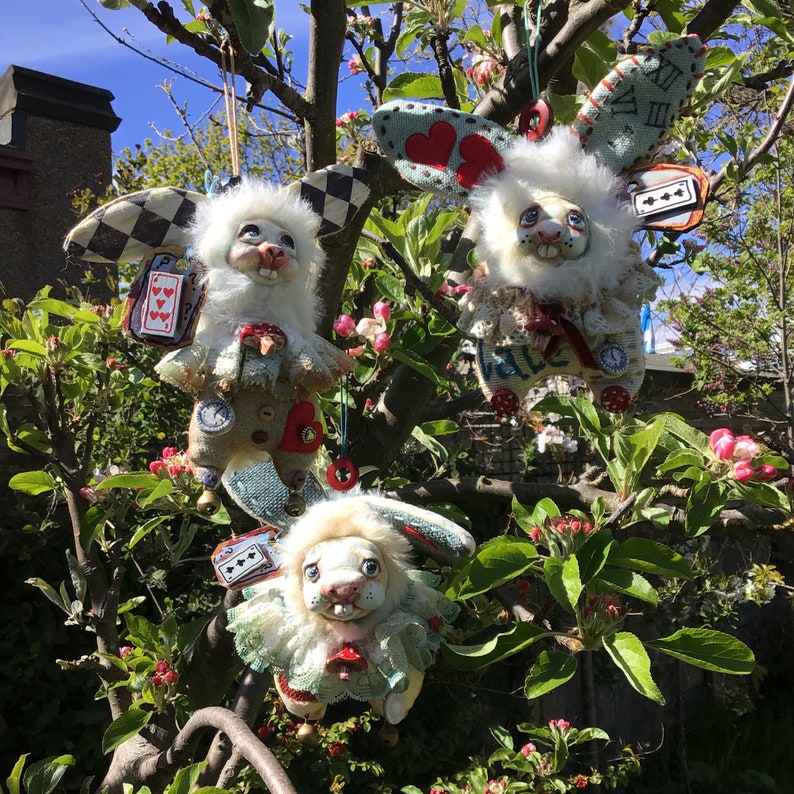 May include: Three white rabbit plushies with different fabric textures and designs. Each plush has a different face and is decorated with fabric flowers, hearts, and playing cards. The plushies are hanging from a tree branch.