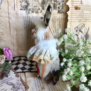May include: A white rabbit doll with a black and white striped hat, a white lace collar, and a red heart on its chest. The doll is sitting on a black and white checkered surface.
