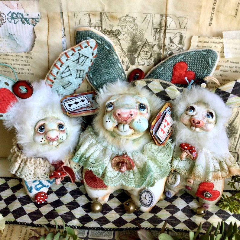 May include: Three white rabbit plushies with painted faces and embroidered details. The plushies are wearing lace collars and have fabric wings with playing card and heart designs. The plushies are sitting on a black and white checkered surface.