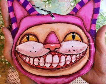 Könnte beinhalten: Eine rosa und violett bemalte Holzkatzenmaske mit einem breiten, zahnigen Grinsen. Die Augen der Katze sind gelb und die Maske hat eine weiße Nase und Schnurrhaare.