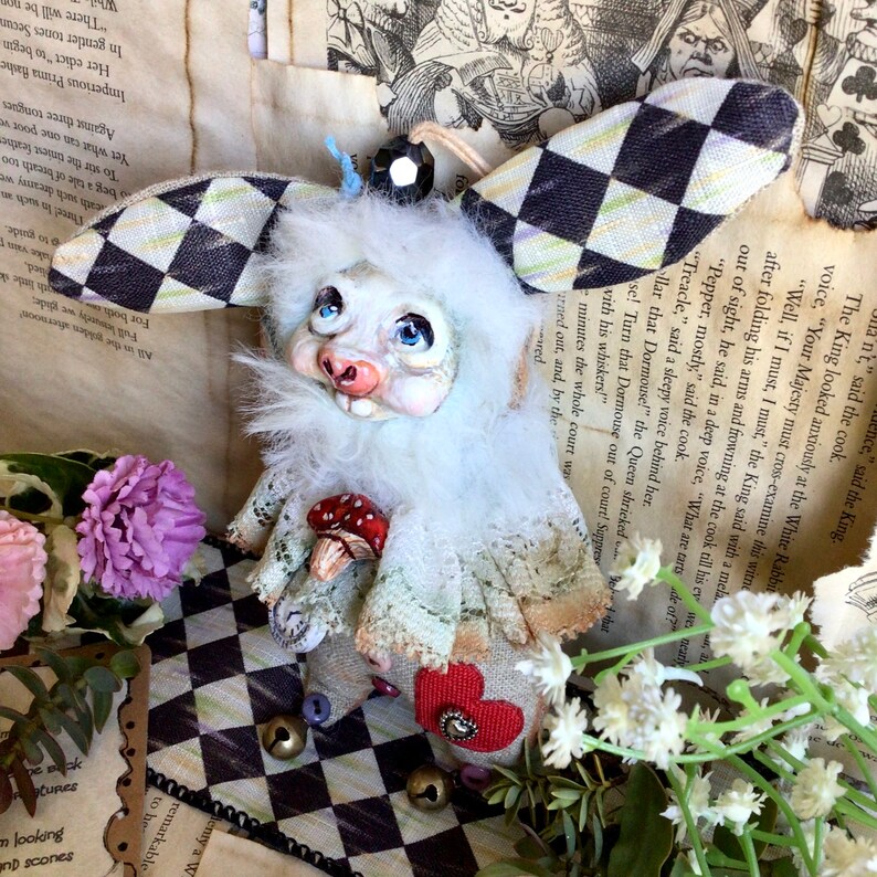 May include: White rabbit plush toy with a checkered fabric hat, holding a red mushroom. The toy has a white fur body, blue eyes, and a pink nose. The toy is sitting on a black and white checkered surface.