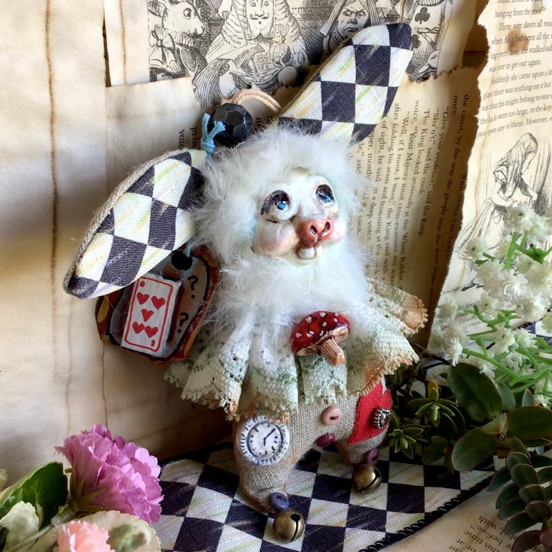 May include: A white rabbit doll with a checkered hat, a lace collar, and a red mushroom. The doll is wearing a brown and white outfit with a clock face on one leg and bells on the other. The doll is sitting on a black and white checkered surface.