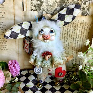 May include: A white rabbit doll with a checkered fabric ear, a red heart, a mushroom, and a pocket watch. The doll is wearing a white lace collar and has a white fur body. The doll is sitting on a black and white checkered surface.