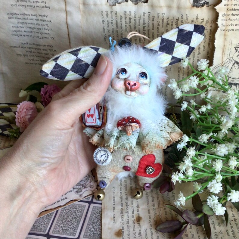 May include: White rabbit plush toy with a black and white checkered fabric ear, a red heart, a clock, and a playing card with the Queen of Hearts. The rabbit is wearing a white lace collar and a mushroom.