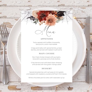 May include: A Halloween-themed menu card on a white plate, with a fork and knife. The menu features a floral arrangement with bats and a spiderweb. The menu includes appetizers, main courses, and desserts.
