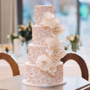 May include: A four-tiered wedding cake with a light pink and white floral design. Large, cream-coloured paper roses cascade down the side. The cake is on a silver cake stand, with a blurred background of a room with tables and chairs.