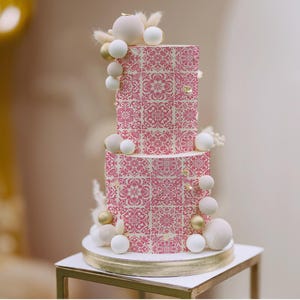May include: A two-tiered cake with a pink and cream floral tile pattern. The cake is decorated with white and gold spherical accents and topped with dried floral elements. It sits on a gold-rimmed white cake stand.