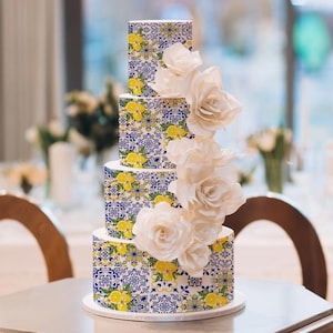 Könnte beinhalten: Ein vierstöckiger Kuchen mit weißer Basis, verziert mit einem blau-gelb-weißen Fliesenmuster. Große weiße Rosen fallen an der Seite herab. Der Kuchen steht auf einem weißen Kuchenständer, ideal für eine Hochzeit oder einen besonderen Anlass.