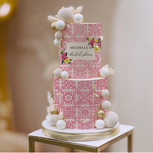 May include: A two-tiered cake with a pink and white floral tile pattern. The cake is decorated with white and gold spheres, and the text "MICHELLE'S bridal shower" is displayed on the top tier.