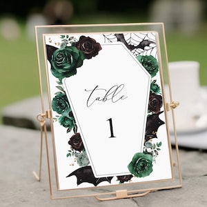 May include: A white table number card with a black coffin shape and the text "Table 1". The card is decorated with green and burgundy roses, black bats, and gold glitter.