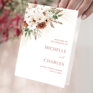 Peut inclure: Un programme de mariage blanc avec des accents floraux crème, marron et verts. Le texte indique "WELCOME TO THE WEDDING OF MICHELLE and CHARLES" avec le lieu et la date en dessous. Le programme est tenu par une personne.