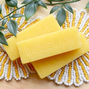May include: Two yellow beeswax blocks on a white and yellow beaded surface with green leafy branches.