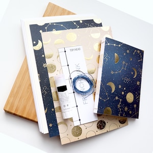 May include: A craft kit for making a journal with a moon and stars theme. The kit includes a stack of paper, a piece of fabric, a glue stick, a piece of string, and a printed guide with the text "TOP/HEAD FRENCHINK".