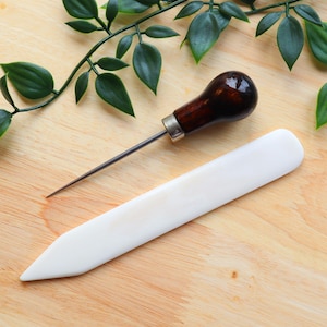 May include: A white leather stitching tool and a brown wood handle awl with a metal tip, both tools are on a wooden surface.