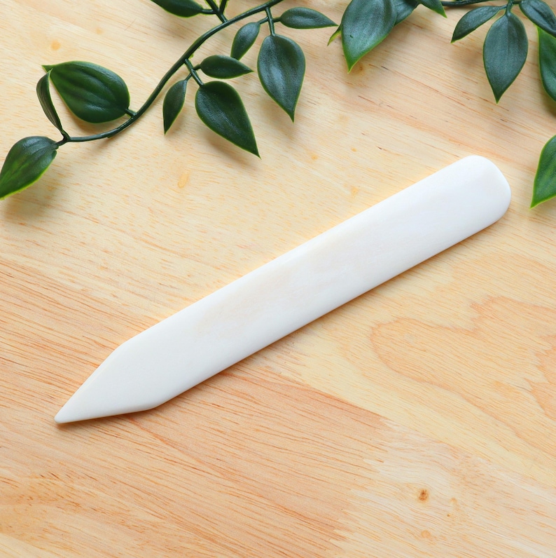 May include: A white bone folder with a pointed tip, lying on a wooden surface. The folder is used for scoring and creasing paper and fabric.