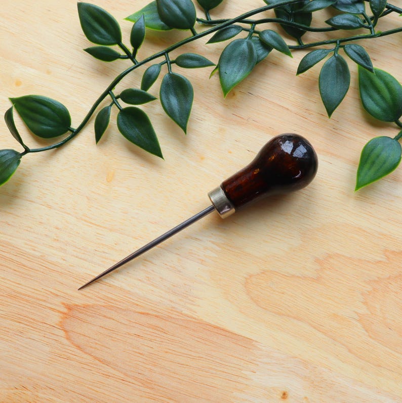 May include: A brown leather crafting tool with a metal needle and a wooden handle. The tool is lying on a wooden surface with green leaves in the background.