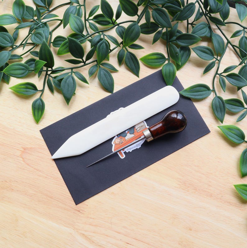 May include: A white leather crafting tool and a brown leather awl with a metal needle on a black background.