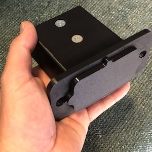 May include: Black plastic mounting bracket with two screws and a cutout for a device. The bracket is held in a hand.