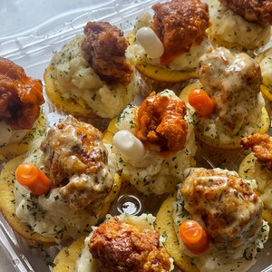 May include: A clear plastic container filled with appetizers. Each appetizer consists of a round, golden-brown base topped with mashed potatoes, fried chicken, and garnishes. Some chicken pieces are coated in a red sauce, and some have a white sauce.
