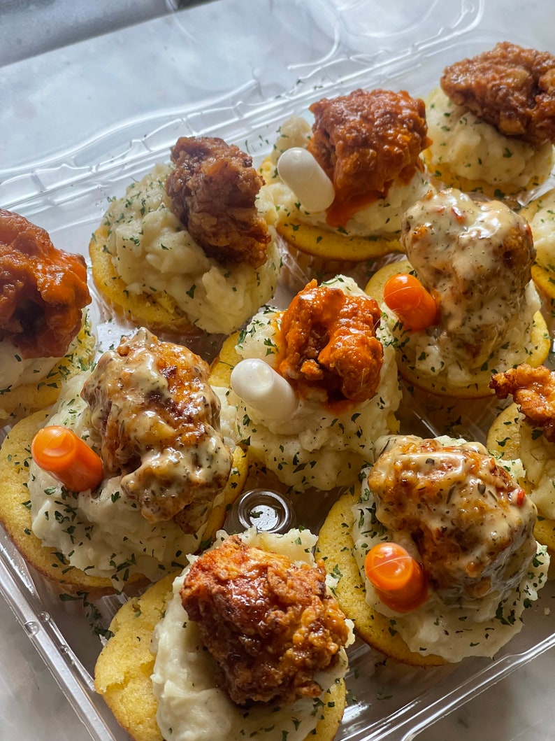 May include: A clear plastic container filled with appetizers. Each appetizer consists of a round, golden-brown base topped with mashed potatoes, fried chicken, and garnishes. Some chicken pieces are coated in a red sauce, and some have a white sauce.