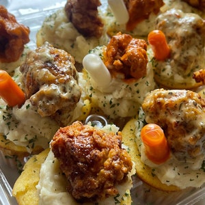 May include: A close-up shot of a tray filled with appetizers. Each appetizer consists of a cornbread base topped with mashed potatoes, fried chicken, and a garnish. Some appetizers have a carrot stick and a white stick as garnish.