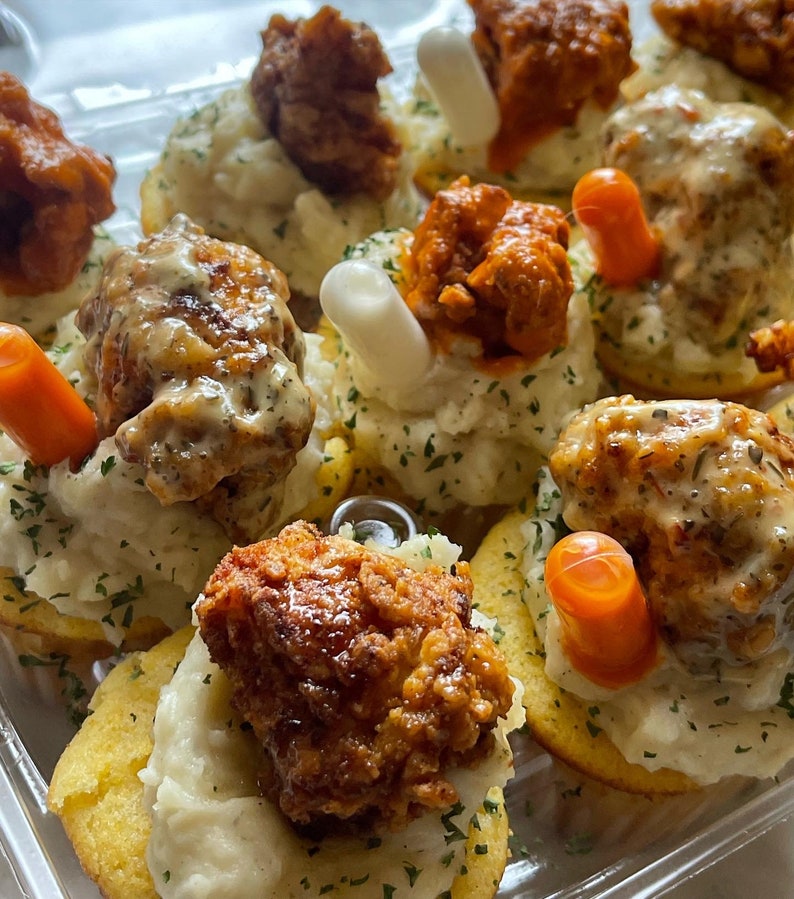 May include: A close-up shot of a tray filled with appetizers. Each appetizer consists of a cornbread base topped with mashed potatoes, fried chicken, and a garnish. Some appetizers have a carrot stick and a white stick as garnish.