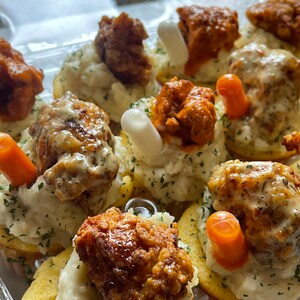 May include: A clear plastic container filled with appetizers. Each appetizer consists of a cornbread base, topped with mashed potatoes, fried chicken, and a garnish of either a carrot stick or a white, cylindrical food item. Some are drizzled with sauce.