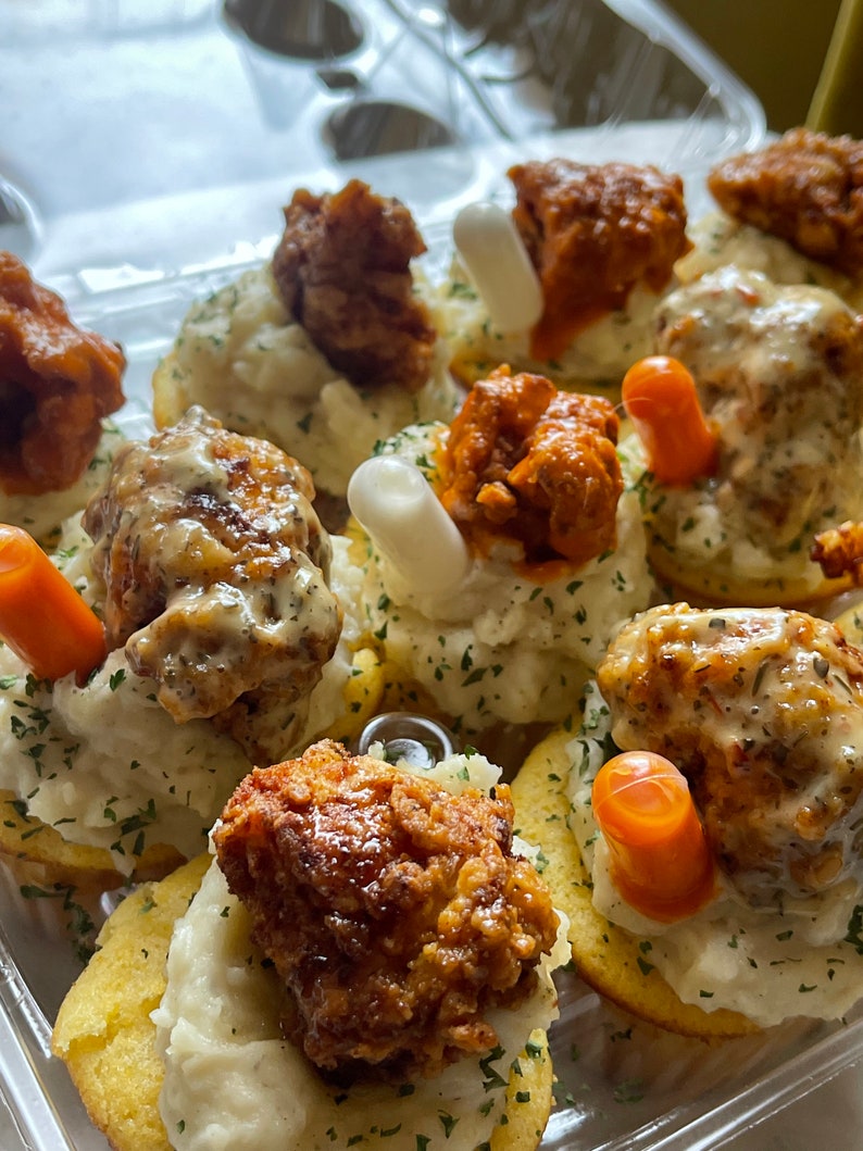 May include: A clear plastic container filled with appetizers. Each appetizer consists of a cornbread base, topped with mashed potatoes, fried chicken, and a garnish of either a carrot stick or a white, cylindrical food item. Some are drizzled with sauce.