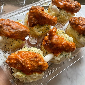 May include: A clear plastic container holds six servings of food. Each serving has a bed of mashed potatoes, a chicken wing coated in orange sauce, and a white, cylindrical garnish. Sprinkled herbs add color.