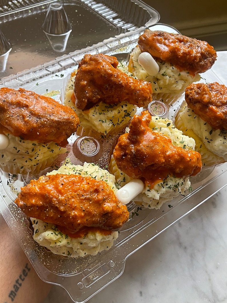 May include: A clear plastic container holds six servings of food. Each serving has a bed of mashed potatoes, a chicken wing coated in orange sauce, and a white, cylindrical garnish. Sprinkled herbs add color.