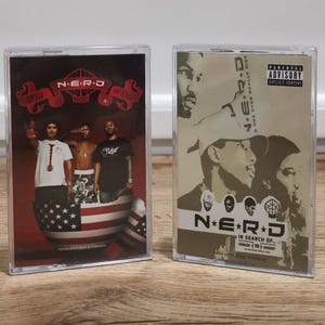 May include: Two cassette tapes featuring the band N.E.R.D. The first tape has a red and black background with the band members and an American flag design. The second tape is tan with band members and the text "N.E.R.D In Search Of..."