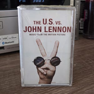 Könnte beinhalten: Eine Kassette mit dem Titel "THE U.S. VS. JOHN LENNON MUSIC FROM THE MOTION PICTURE." Das Cover zeigt eine Hand, die ein Friedenszeichen formt, mit Sonnenbrillen im Design der amerikanischen Flagge.