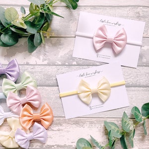 May include: Assortment of pastel satin hair bows with matching elastic headbands. The bows are arranged in a rainbow pattern, with colours including light pink, lavender, yellow, peach, and green. The headbands are white.