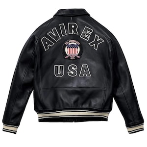 May include: Black leather jacket with the word "AVIREX" arched across the shoulders, "USA" below, and a circular patch with the American flag. The jacket has a ribbed collar, cuffs, and hem with white stripes.