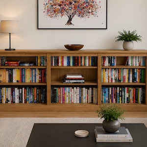 May include: A light oak bookcase filled with books of various colours. The bookcase has three sections and sits against a white wall. A painting of a colourful tree hangs above the bookcase. A lamp and potted plants are also visible.