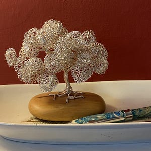 May include: A silver wire tree sculpture on a wooden base, displayed in a white ceramic dish. A patterned pen with blue and green swirls rests in the dish. The background is a solid red.