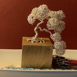 Op de afbeelding: Zilveren draad bonsai boom sculptuur op een houten blok. De boom heeft een gedraaide stam en een volle, getextureerde kroon. Een rode en zilveren aansteker en een pen met een blauw en groen patroon staan op de voorgrond.